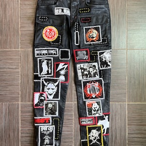 May include: Black denim jeans with various patches sewn on. Patches include a skull and crossbones, a demon face, a band logo, and a lightning bolt. The jeans have a distressed look.