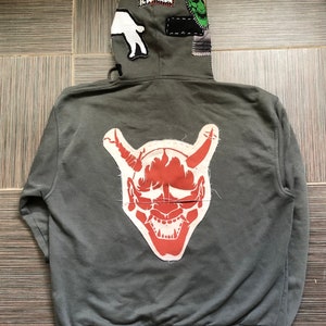 Puede incluir: Una sudadera con capucha gris con un parche de máscara de demonio japonés rojo y blanco en la espalda. La capucha tiene varios parches cosidos, incluido un parche blanco con un contorno negro de una persona.