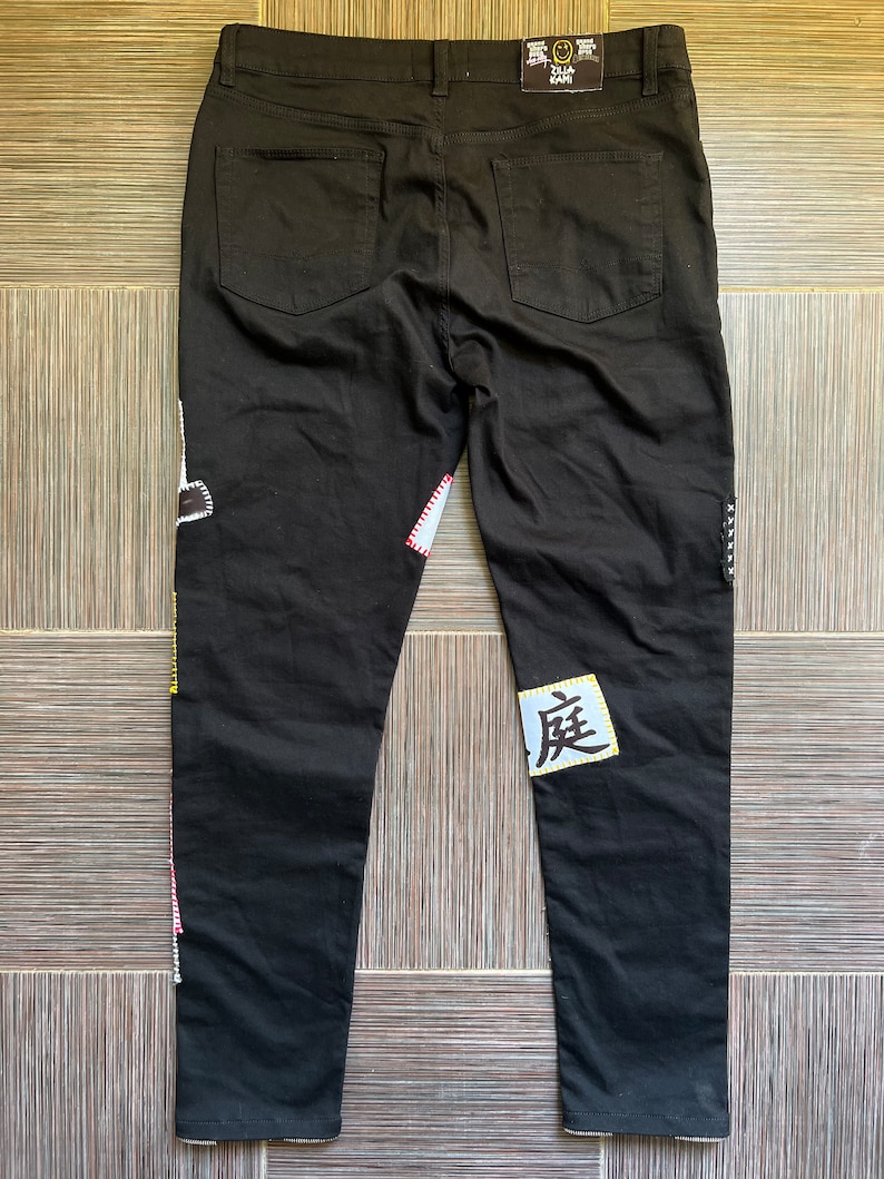 May include: Black jeans with colourful patches and a white patch with the Japanese character "度" on the right leg. The jeans have a black tag with the text "ZILLA KAMI" on the back.