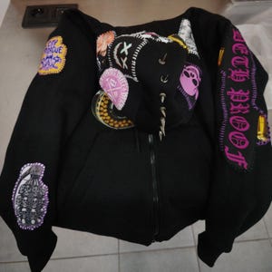May include: Black zip-up hoodie with multiple patches and embellishments. Patches include text, skulls, and other graphics in pink, yellow, and white. The hood features silver spikes. The right sleeve has the text "COLD PROOF" in pink.