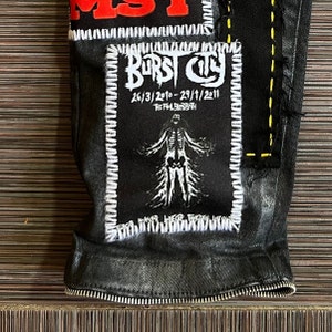 May include: Black denim jacket with a white and yellow stitched patch that says "BURST CITY 26/3/2010-29/1/2011 THE FINAL BLOODBATH" and a white skeleton graphic.