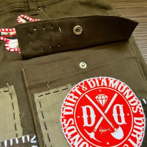 May include: Close-up of a pair of olive green cargo pants with a red and white patch that says "Dirt & Diamonds" with a crossed shovel and pickaxe design. The pants have multiple pockets and are made of a durable fabric.