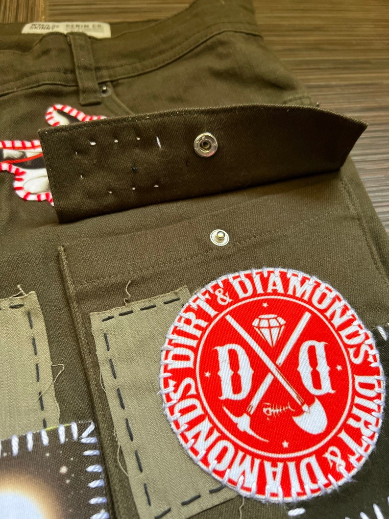 May include: Close-up of a pair of olive green cargo pants with a red and white patch that says "Dirt & Diamonds" with a crossed shovel and pickaxe design. The pants have multiple pockets and are made of a durable fabric.