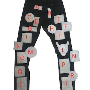 May include: Black denim jeans with various white and silver patches and stickers. The patches are in the shape of circles, squares, and stars. The stickers have letters from A to T on them.