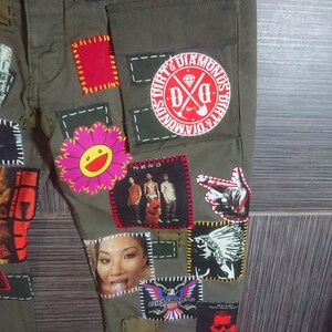 May include: Green cargo pants with various patches sewn on. Patches include a flower, a band logo, a cartoon face, and a graphic of a Native American. The pants have a large pocket on the right leg.