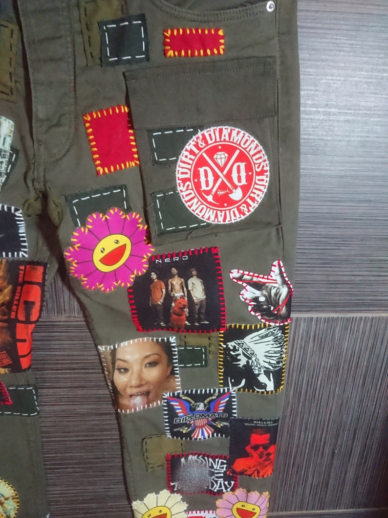 May include: Green cargo pants with various patches sewn on. Patches include a flower, a band logo, a cartoon face, and a graphic of a Native American. The pants have a large pocket on the right leg.