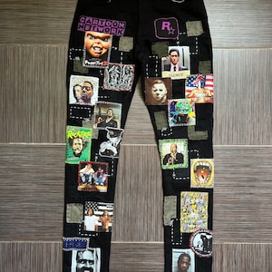 May include: Black denim jeans with various patches sewn on. The patches feature images of characters from movies and TV shows, including a clown, a man with a knife, and a man with a mask. The text "Cartoon Network" and "Oldboy" are visible on the patches.