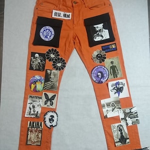 May include: Orange denim jeans with various patches and embroidered designs, including a smiley face, a butterfly, and a wolf. The jeans have black zippers at the bottom of the legs and a black pocket on the right leg with a graphic of a person wearing a hat. The text "使徒、殲滅" is written on a white patch on the front of the jeans.