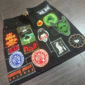 Op de afbeelding: Zwarte denim rok met verschillende opgenaaide patches, waaronder een groen en wit demonen gezicht, een rood en wit schedel en gekruiste beenderen, een gele en rode smiley en een zwarte en witte bloem. De tekst "DIRT & DIAMONDS" wordt op verschillende patches herhaald.