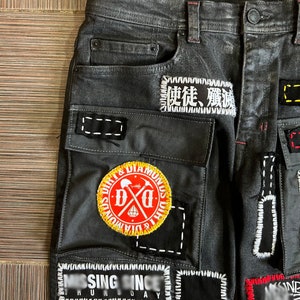 May include: Black denim jeans with multiple patches sewn on. Patches include a skull and crossbones with the text "Dirt & Diamonds", a patch with Japanese characters, and a patch with the text "Missing Since" and "Humanity Day".