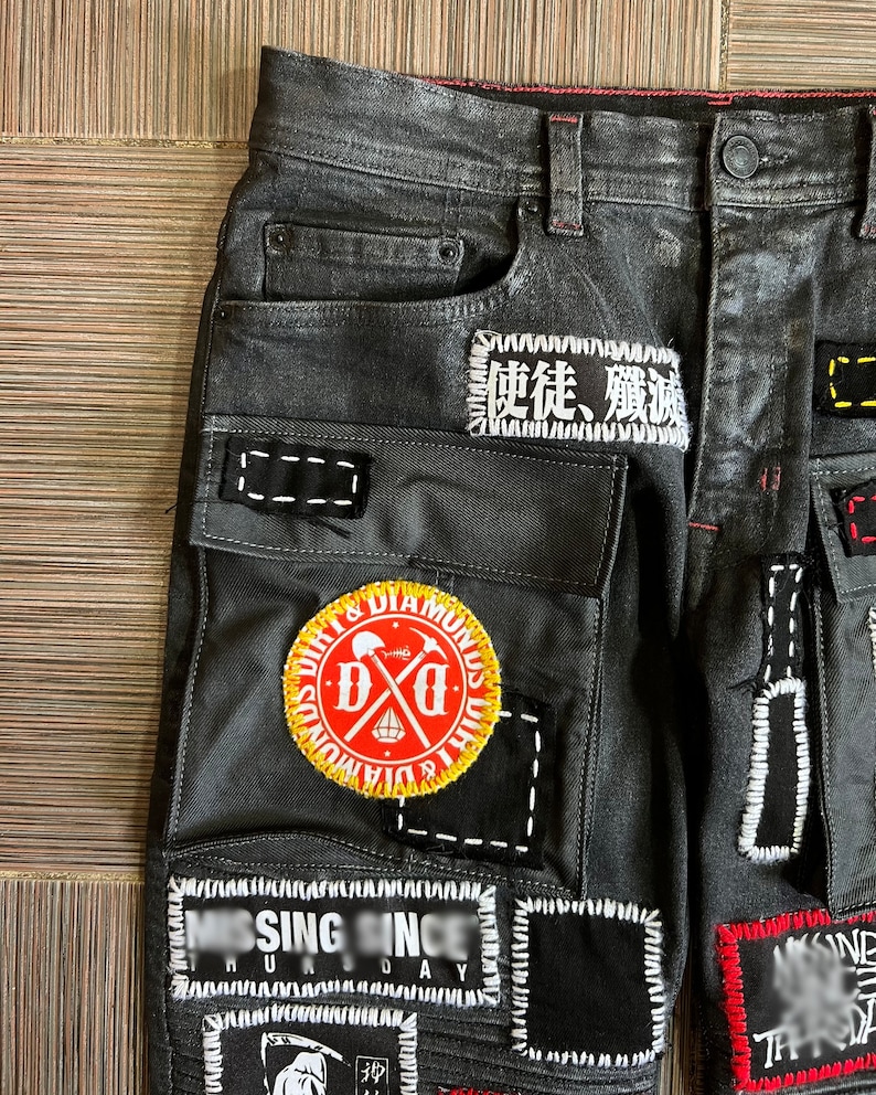 May include: Black denim jeans with multiple patches sewn on. Patches include a skull and crossbones with the text "Dirt & Diamonds", a patch with Japanese characters, and a patch with the text "Missing Since" and "Humanity Day".