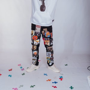 May include: Black jeans with a white hoodie and a variety of colorful patches and stickers. The patches include images of people, logos, and text. The jeans are worn by a person standing on a white background.