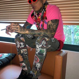 May include: A person wearing a pink t-shirt with a colorful scarf, black sunglasses, and green patched jeans. The person is sitting on a brown couch.