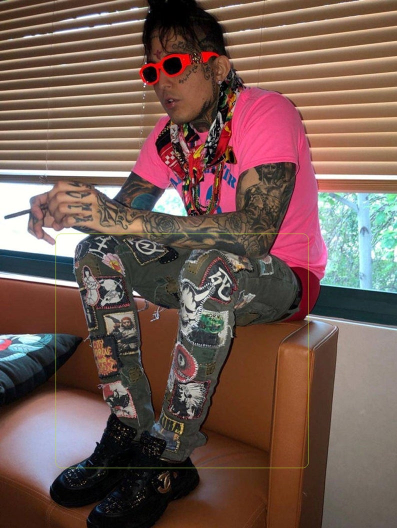 May include: A person wearing a pink t-shirt with a colorful scarf, black sunglasses, and green patched jeans. The person is sitting on a brown couch.