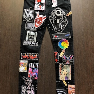 Può includere: Jeans in denim nero con vari patch e disegni, tra cui un teschio, una pistola, una faccina sorridente e un personaggio dei cartoni animati. I jeans hanno un aspetto consumato e sono ricoperti di cuciture.