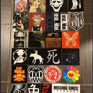 May include: A collection of stickers featuring various designs, including a demon mask, a butterfly, a skull, a bear, a flower, and a city morgue logo. The stickers are arranged in a grid pattern on a brown tiled surface.