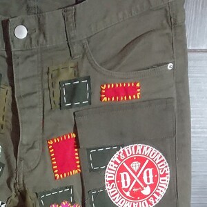 May include: Green cargo pants with multiple patches sewn on. The patches are red, yellow, and white. One patch is a circle with the text "Dirt & Diamonds" and a crossed shovel and pickaxe.