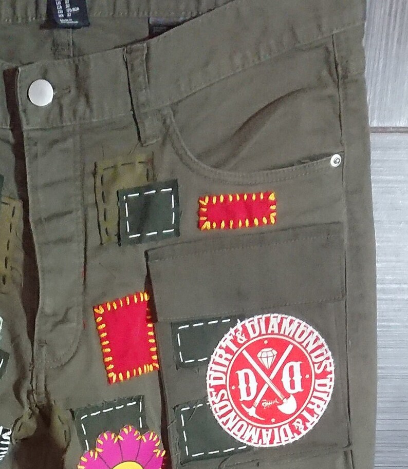May include: Green cargo pants with multiple patches sewn on. The patches are red, yellow, and white. One patch is a circle with the text "Dirt & Diamonds" and a crossed shovel and pickaxe.