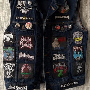 May include: A dark blue denim vest featuring numerous embroidered band patches and circular pinback buttons. The patches display various graphic designs and band names including Nasty Savage, Judas Priest, Entombed, and Obituary. A Wrangler brand label is visible.