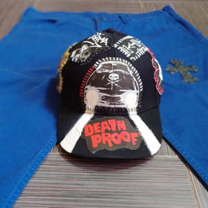 May include: A black baseball cap with various patches, including a skull and crossbones, a car, and the words "Death Proof".
