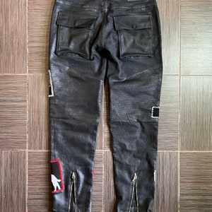 May include: Black denim pants with leather patch pockets and zipper details on the legs. The pants have a distressed look and feature a white graphic on the left leg.