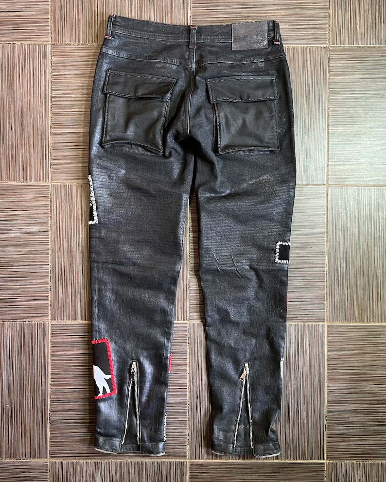 May include: Black denim pants with leather patch pockets and zipper details on the legs. The pants have a distressed look and feature a white graphic on the left leg.