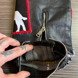 May include: Black denim jeans with a white figure patch and a silver zipper. The zipper is partially open, revealing the inside of the jeans.