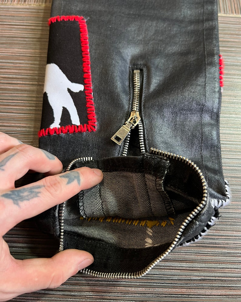May include: Black denim jeans with a white figure patch and a silver zipper. The zipper is partially open, revealing the inside of the jeans.