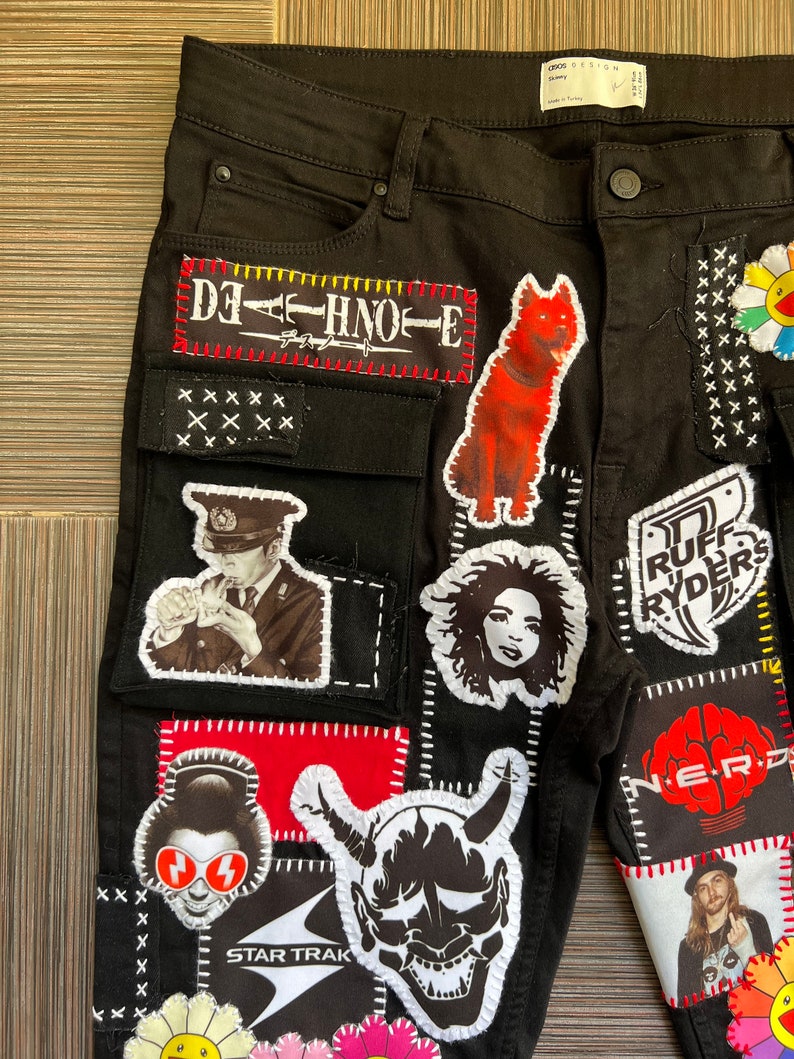 May include: Black denim jeans with various patches sewn on, including a red dog, a black and white portrait of a man in a hat, a red and white demon, a black and white portrait of a woman, a red and white flower, and a black and white logo with the text "STAR TRAK".