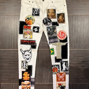 Puede incluir: Vaqueros blancos de denim con varios parches y diseños, incluyendo una calavera, una mariposa, un demonio y una cara de mujer. Los vaqueros tienen detalles desgastados y son una prenda única y llamativa.