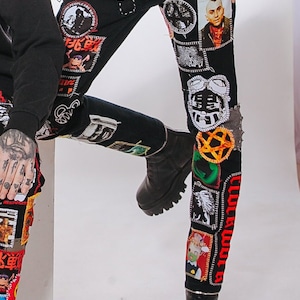 Puede incluir: Vaqueros negros de denim con varios parches y diseños, incluyendo una calavera, un retrato y una estrella. Los vaqueros tienen un cinturón con tachuelas plateadas.