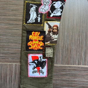 May include: Green cargo pants with various patches sewn on. Patches include a skull and crossbones, a woman in bondage, a grim reaper, a dog, and a person holding a gun. The text "City Morgue" and "Hikari Ultra" are visible on the patches.