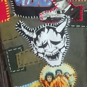 May include: A green fabric jacket with various patches sewn on. One patch features a black and white illustration of a Japanese demon mask with horns. Another patch features a band logo with the word "WIND" in blue and white letters.