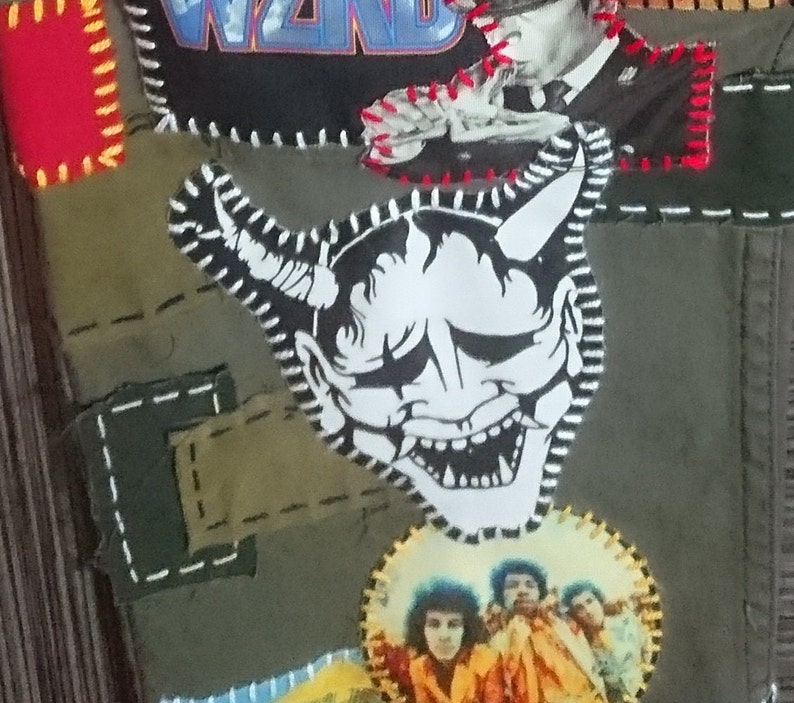 May include: A green fabric jacket with various patches sewn on. One patch features a black and white illustration of a Japanese demon mask with horns. Another patch features a band logo with the word "WIND" in blue and white letters.