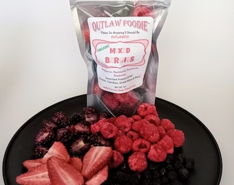 May include: A bag of freeze-dried mixed berries, including strawberries, raspberries, blackberries, and blueberries. The bag is labeled "Outlaw Foodie" and "Mixed Berries". The berries are arranged on a black plate.