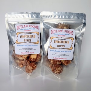 Può includere: Due buste metalliche argentate contenenti bocconcini di Snickers liofilizzati. Ogni etichetta riporta il testo Outlaw Foodie, Flavor So Amazing It Should Be Outlawed!, Freeze Dried Snickers Bites, We Take Snacking To The Next Level! e OutLawFoodie.etsy.com.