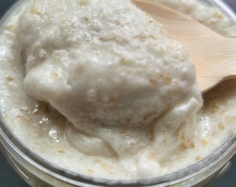 May include: A close-up of a white, creamy, and grainy scrub in a clear jar. A wooden spoon is scooping out the scrub.