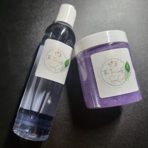 May include: A clear bottle and a jar of purple body scrub. Both containers have white labels with the text "The Full Lady Soap Co." and a peacock feather design. The bottle has a white cap.