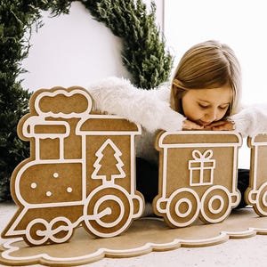 Könnte beinhalten: Ein Weihnachts-Zug aus Holz mit Lebkuchen-Optik. Die Waggons zeigen einen Weihnachtsbaum und ein Geschenkbox-Design, weiß umrandet. Der Zug steht auf einer dekorativen Basis mit gewelltem Rand.