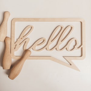 Puede incluir: Un letrero de madera con forma de burbuja de diálogo con la palabra "hello" recortada. El letrero está rodeado por tres bolos de madera.