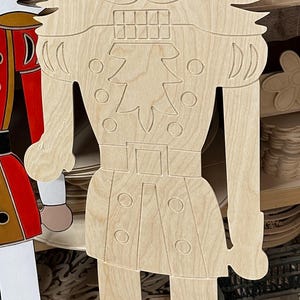Op de afbeelding: Een grote, onafgewerkte houten uitsparing van een notenkrakersoldaat. De uitsparing heeft een kroon, gedetailleerde gelaatstrekken, een uniform met knopen en laarzen. Het ontwerp is in het hout gesneden en toont de natuurlijke nerf.