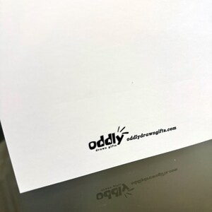 May include: A white card with the black text "oddly drawn gifts" and the website oddlydrawngifts.com. The card is angled, with a reflection of the text visible below.