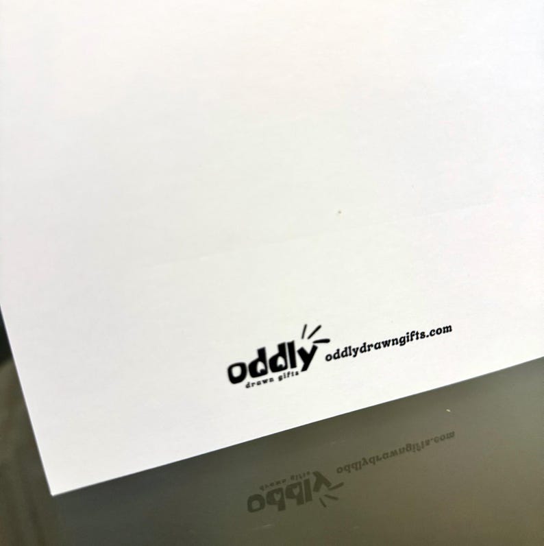 May include: A white card with the black text "oddly drawn gifts" and the website oddlydrawngifts.com. The card is angled, with a reflection of the text visible below.