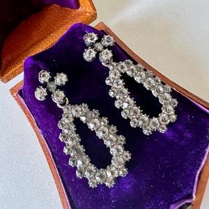 May include: A pair of antique silver earrings with teardrop-shaped designs, set with sparkling clear stones, displayed in a purple velvet-lined box.