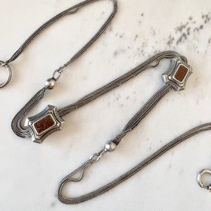 May include: A silver chain with two decorative elements. Each element has a rectangular shape with a brown stone in the center.