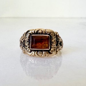 May include: A gold ring with an ornate design and a square amber stone in the centre.