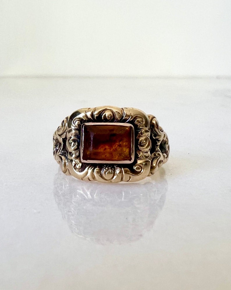 May include: A gold ring with an ornate design and a square amber stone in the centre.