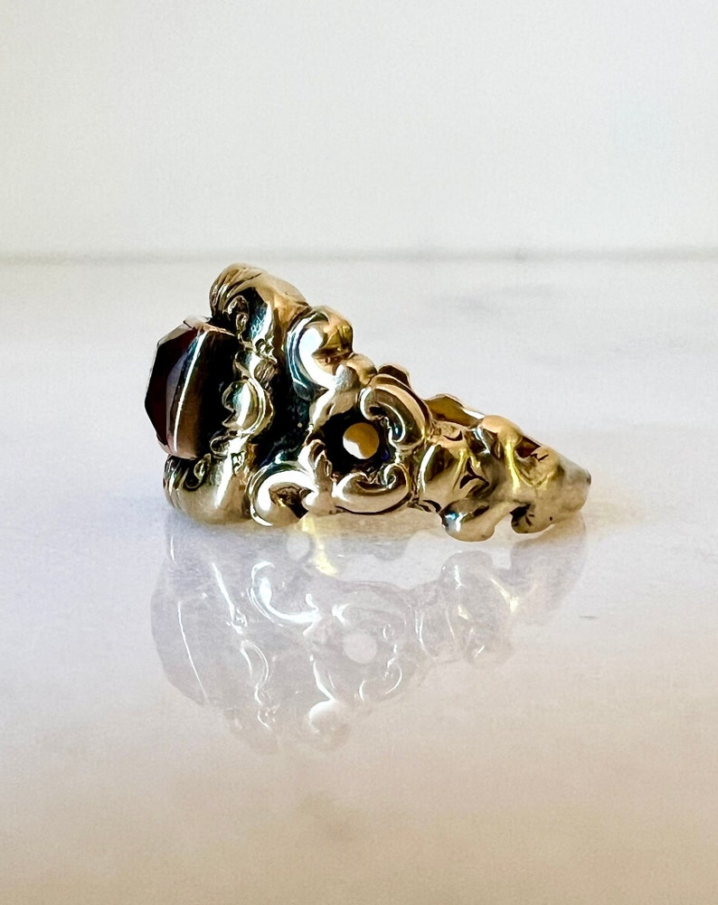 May include: A gold ring featuring a large, dark red gemstone set within an intricate, ornate design.
