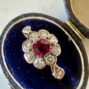 May include: A vintage ring featuring a red ruby surrounded by a halo of small diamonds. The ring is set in silver and is presented in a blue velvet ring box.
