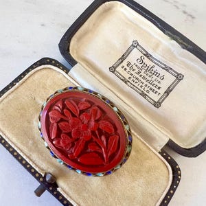 May include: A red carved floral brooch with a blue and white enamel border, in a black velvet box with a white label that reads "Spink's From Dent Ltd. The Jewellers 28 Church Street Enfield."